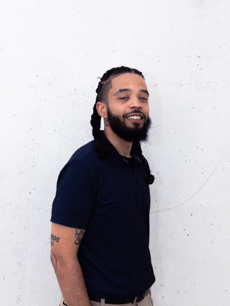 Portrait of Khalil Henry, a Saint Louis real estate agent with SoldWithKhalil.com, standing confidently against a white wall, wearing a navy polo shirt with braided hair and visible tattoos on his arm.